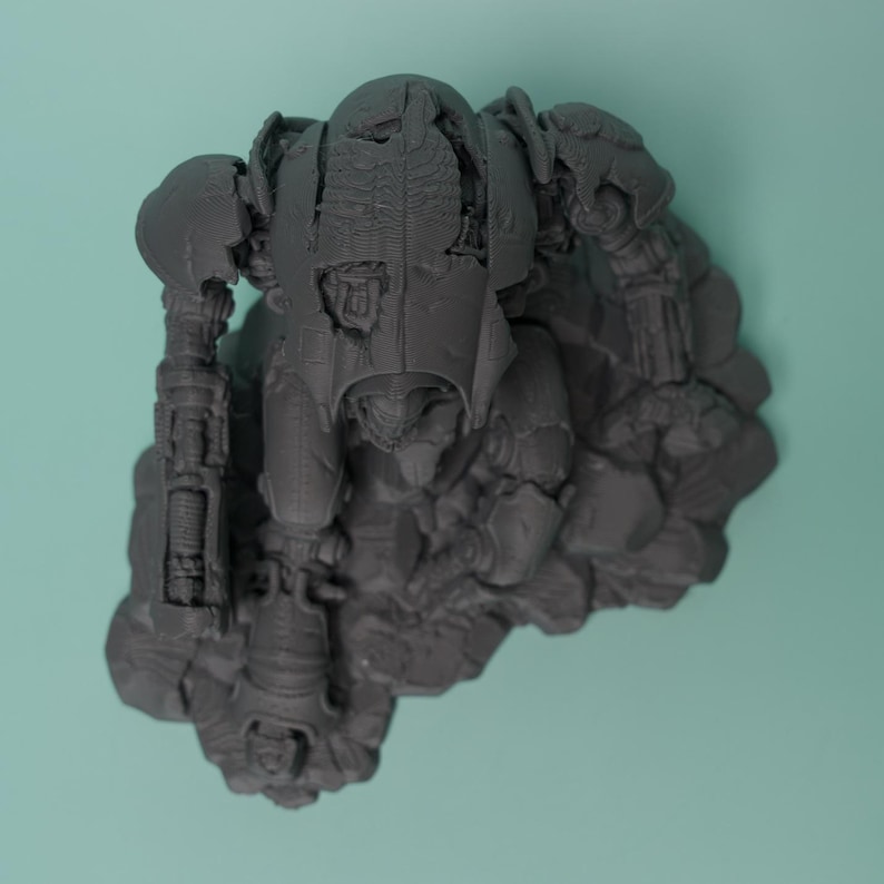 May include: A dark gray, 3D-printed miniature robot figure in a kneeling position, featuring intricate mechanical details, articulated limbs, and a textured armor plating surface, mounted on a rocky, uneven base with visible layer lines.