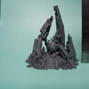 Może przedstawiać: Szary, wydrukowany w 3D miniaturowy element terenu przedstawiający centralną, pękniętą kulę otoczoną poszarpanymi, kanciastymi ruinami i gruzem. Metalowa linijka umieszczona pionowo obok konstrukcji wskazuje wysokość około 20 cm.