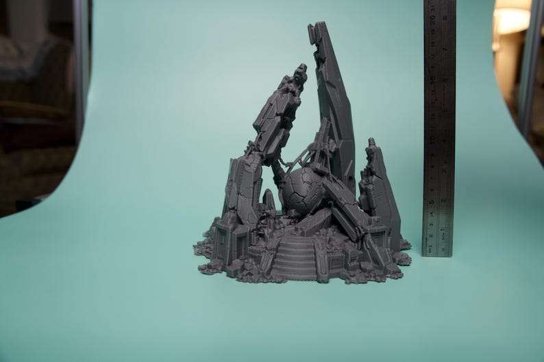 Może przedstawiać: Szary, wydrukowany w 3D miniaturowy element terenu przedstawiający centralną, pękniętą kulę otoczoną poszarpanymi, kanciastymi ruinami i gruzem. Metalowa linijka umieszczona pionowo obok konstrukcji wskazuje wysokość około 20 cm.