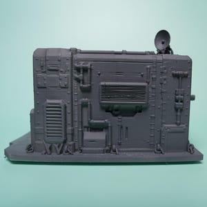 May include: A gray, rectangular, industrial-style miniature terrain piece featuring intricate molded details such as pipes, vents, a rectangular grille, and a small satellite dish mounted on the top right corner, all set on a flat base.