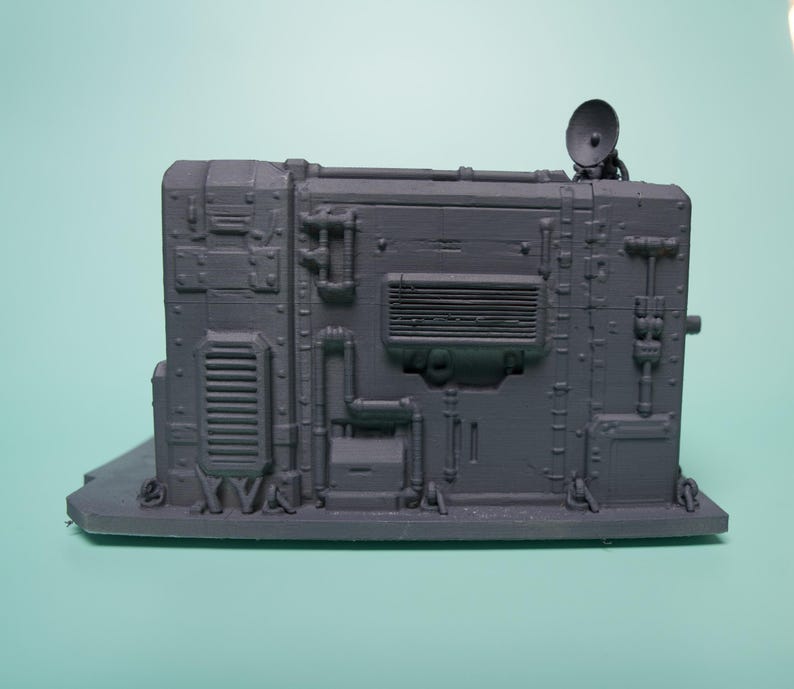 May include: A gray, rectangular, industrial-style miniature terrain piece featuring intricate molded details such as pipes, vents, a rectangular grille, and a small satellite dish mounted on the top right corner, all set on a flat base.