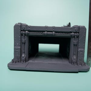 May include: A gray, unpainted plastic miniature terrain piece representing a sci-fi hangar or bunker entrance with industrial detailing, rivets, and structural beams. A metal ruler is placed vertically next to it, showing a height of 6 inches.