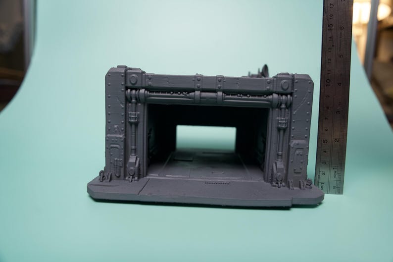 May include: A gray, unpainted plastic miniature terrain piece representing a sci-fi hangar or bunker entrance with industrial detailing, rivets, and structural beams. A metal ruler is placed vertically next to it, showing a height of 6 inches.