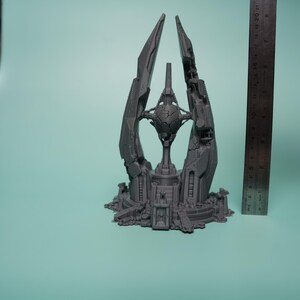 K&ouml;nnte beinhalten: Eine graue 3D-gedruckte Skulptur einer futuristischen Turmstruktur mit einer zentralen Kugel, platziert neben einem Metalllineal, das eine H&ouml;he von etwa 20 cm anzeigt. Das Objekt weist komplexe geometrische Paneele und eine gestufte Basis auf.