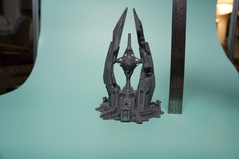 K&ouml;nnte beinhalten: Eine graue 3D-gedruckte Skulptur einer futuristischen Turmstruktur mit einer zentralen Kugel, platziert neben einem Metalllineal, das eine H&ouml;he von etwa 20 cm anzeigt. Das Objekt weist komplexe geometrische Paneele und eine gestufte Basis auf.