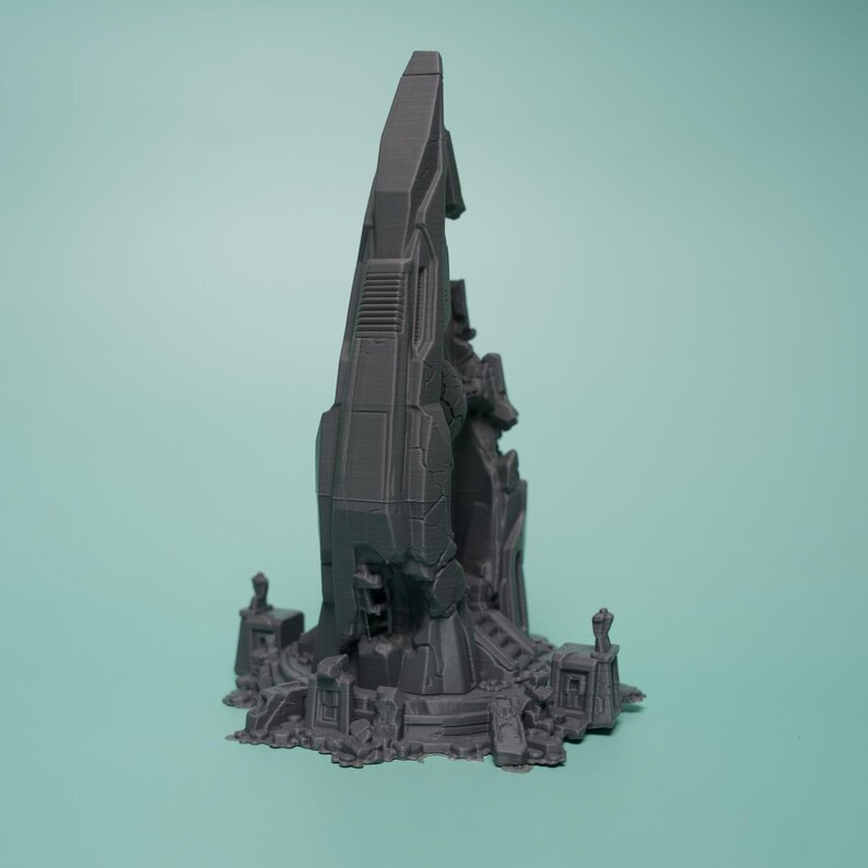 K&ouml;nnte beinhalten: Ein dunkelgraues, 3D-gedrucktes Miniatur-Gel&auml;ndest&uuml;ck, das einen hohen, gezackten, monolithischen Turm mit mechanischen Paneelen und geometrischen Details zeigt, platziert auf einer Basis mit kleineren architektonischen Ruinen und verstreuten Tr&uuml;mmern.