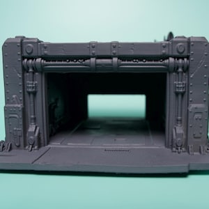 May include: A gray 3D printed miniature terrain piece featuring a rectangular bunker entrance with industrial detailing, including vertical pipes, rivets, and a flat base. The structure has a hollow interior and a wide, open doorway.