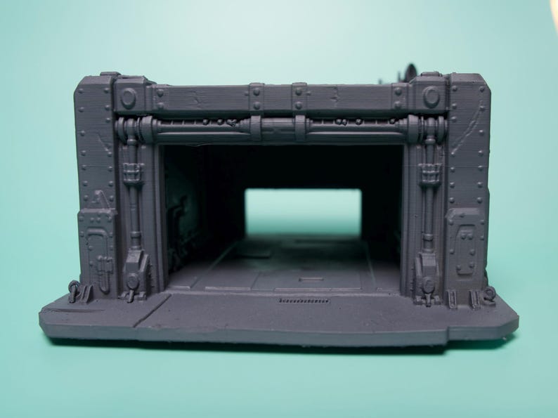May include: A gray 3D printed miniature terrain piece featuring a rectangular bunker entrance with industrial detailing, including vertical pipes, rivets, and a flat base. The structure has a hollow interior and a wide, open doorway.