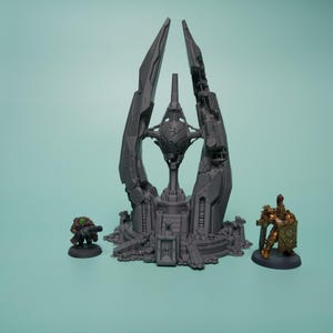 K&ouml;nnte beinhalten: Eine graue, unbemalte 3D-gedruckte Sci-Fi-Gel&auml;ndestruktur mit einer zentralen Kugel, die zwischen hohen, gezackten T&uuml;rmen schwebt. Flankiert wird das Bauwerk von zwei Miniaturfiguren: einer kleinen gr&uuml;n gekleideten Figur und einem goldgepanzerten Krieger.