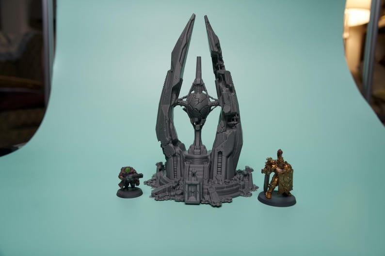 K&ouml;nnte beinhalten: Eine graue, unbemalte 3D-gedruckte Sci-Fi-Gel&auml;ndestruktur mit einer zentralen Kugel, die zwischen hohen, gezackten T&uuml;rmen schwebt. Flankiert wird das Bauwerk von zwei Miniaturfiguren: einer kleinen gr&uuml;n gekleideten Figur und einem goldgepanzerten Krieger.