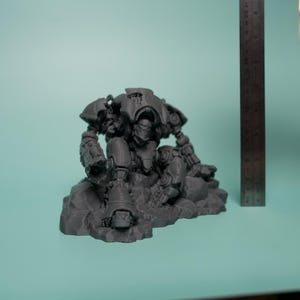 May include: A dark gray, unpainted resin miniature of a mechanical robot figure in a seated position on a rocky base. The figure is positioned next to a metal ruler showing a height of approximately 3.5 inches from the base to the top of the figure.