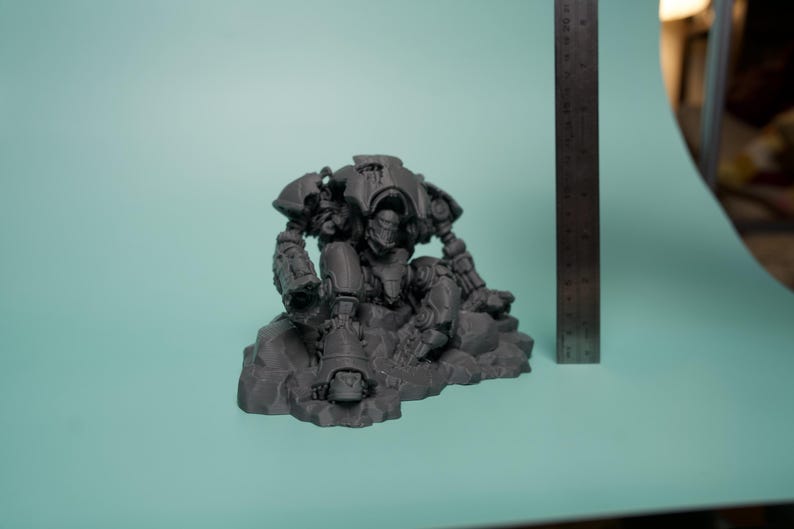 May include: A dark gray, unpainted resin miniature of a mechanical robot figure in a seated position on a rocky base. The figure is positioned next to a metal ruler showing a height of approximately 3.5 inches from the base to the top of the figure.