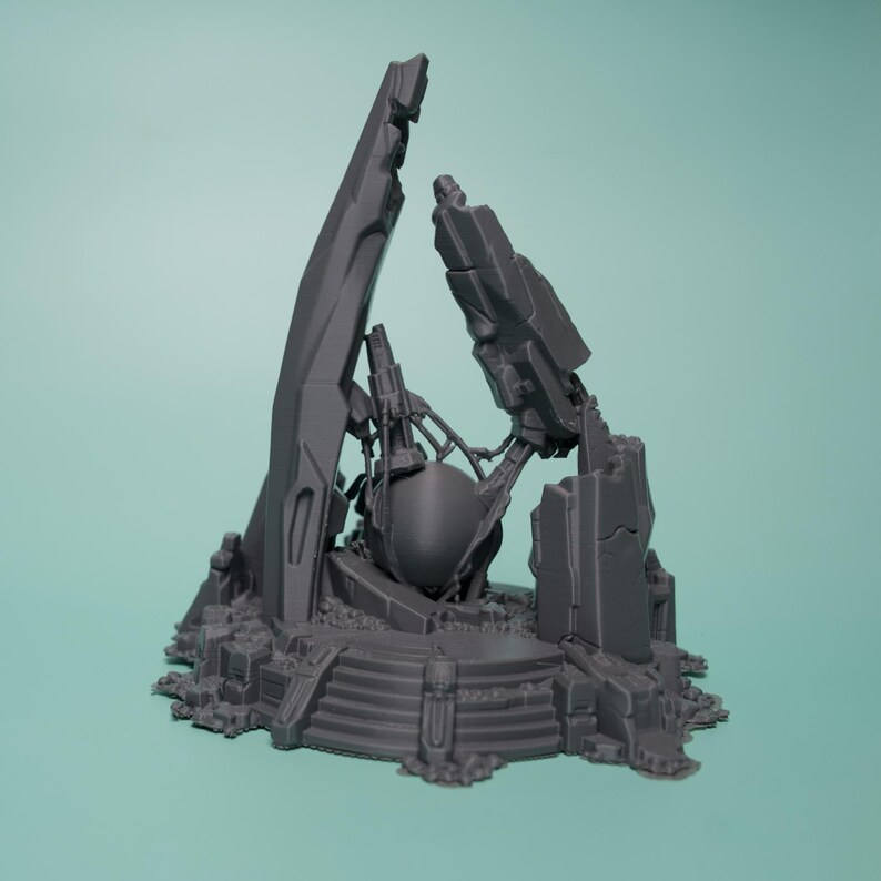 Może przedstawiać: Ciemnoszary, wydrukowany w 3D miniaturowy element terenu przedstawiający centralny sferyczny obiekt zamknięty w poszarpanych, zrujnowanych strukturach architektonicznych i gruzach, umieszczony na okrągłej podstawie ze schodkami i skalistym podłożem.