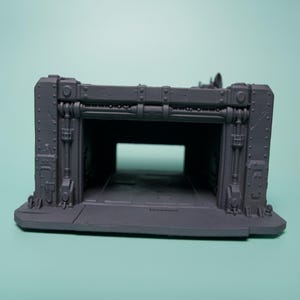 May include: A gray, 3D-printed miniature structure featuring a rectangular hangar-style opening. The exterior walls display industrial details including vertical pipes, rivets, and reinforced paneling, creating a modular terrain piece.