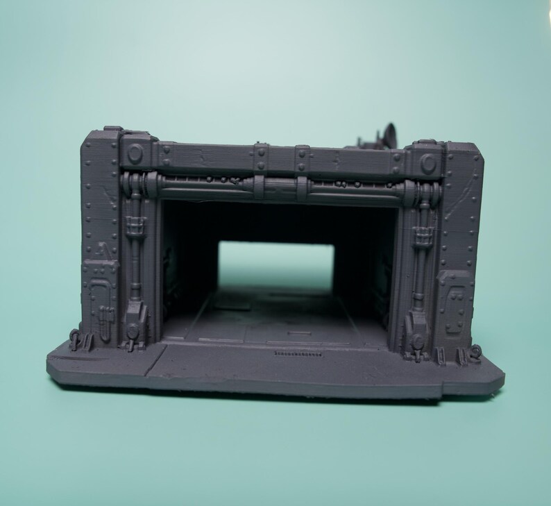 May include: A gray, 3D-printed miniature structure featuring a rectangular hangar-style opening. The exterior walls display industrial details including vertical pipes, rivets, and reinforced paneling, creating a modular terrain piece.
