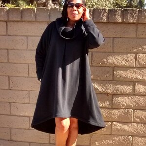 May include: A black, oversized dress with a cowl neck and long sleeves. The dress has a flared, asymmetrical hemline. The person is wearing black open-toe booties. The background is a brick wall.