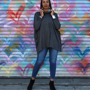 May include: A woman wearing a dark gray cowl-neck sweater, blue jeans, and black ankle boots. The background is a colorful mural with various shades of pink, blue, yellow, and purple. The sweater has long sleeves and a loose fit.