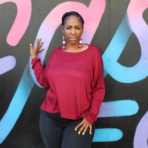 May include: A burgundy long-sleeve top with a relaxed fit. The top is paired with dark-wash jeans. The person is wearing large, colorful earrings and has yellow nail polish. The background is a black wall with colorful graffiti.