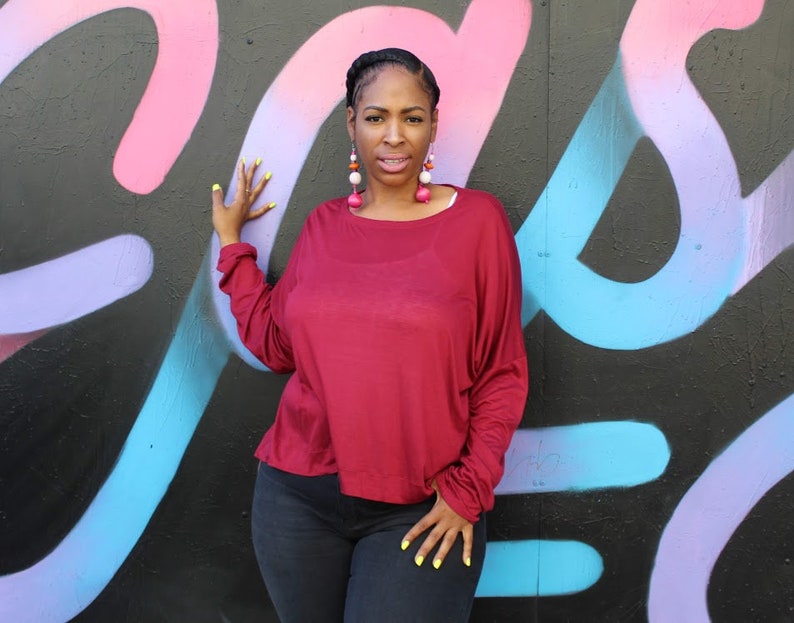 May include: A burgundy long-sleeve top with a relaxed fit. The top is paired with dark-wash jeans. The person is wearing large, colorful earrings and has yellow nail polish. The background is a black wall with colorful graffiti.