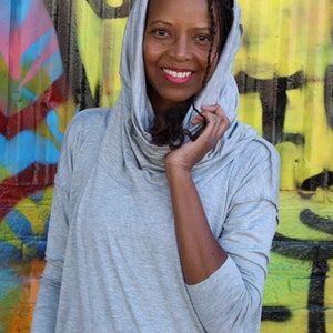 May include: A light gray hooded top with a cowl neck. The top is made of a soft material and is being worn in front of a colorful graffiti wall. The person wearing the top is smiling.