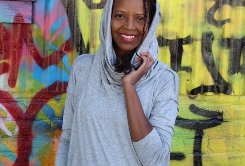 May include: A light gray hooded top with a cowl neck. The top is made of a soft material and is being worn in front of a colorful graffiti wall. The person wearing the top is smiling.