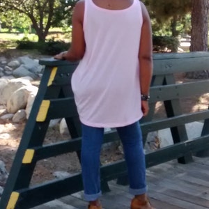 May include: A light pink tank top paired with blue jeans and brown heeled boots. The tank top has a rounded neckline and a loose fit. The jeans are cuffed at the ankles. The boots have a block heel.
