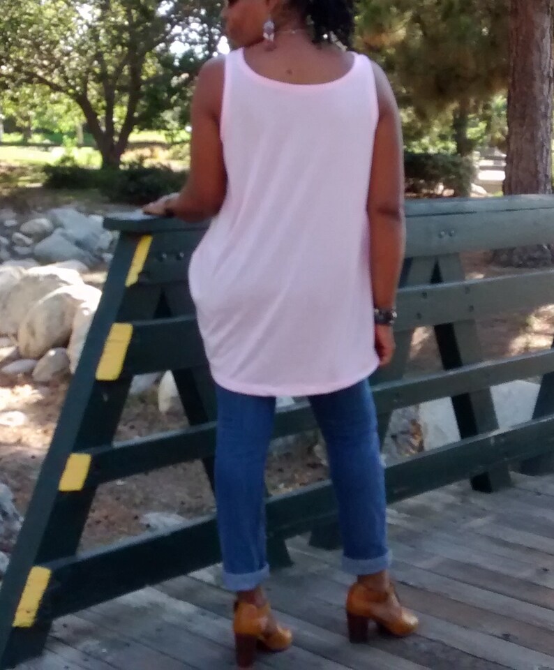 May include: A light pink tank top paired with blue jeans and brown heeled boots. The tank top has a rounded neckline and a loose fit. The jeans are cuffed at the ankles. The boots have a block heel.
