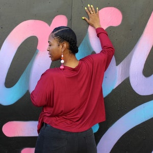 May include: A burgundy long-sleeve top with a draped neckline and side tie. The person is wearing large, dangling earrings with pink, white, and orange beads. The background is a black wall with colorful graffiti in pink, purple, and blue.