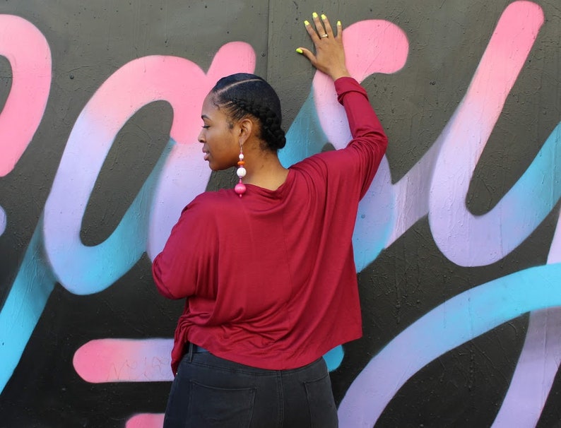 May include: A burgundy long-sleeve top with a draped neckline and side tie. The person is wearing large, dangling earrings with pink, white, and orange beads. The background is a black wall with colorful graffiti in pink, purple, and blue.