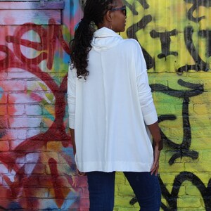 May include: A white long-sleeve tunic with a cowl neck. The tunic is paired with dark blue jeans. The background features a colorful graffiti wall with red, yellow, and black paint.