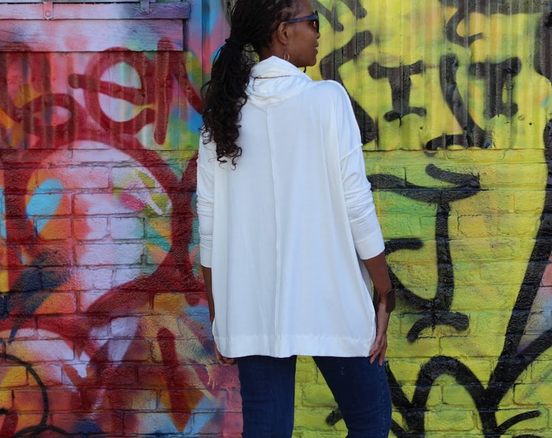 May include: A white long-sleeve tunic with a cowl neck. The tunic is paired with dark blue jeans. The background features a colorful graffiti wall with red, yellow, and black paint.