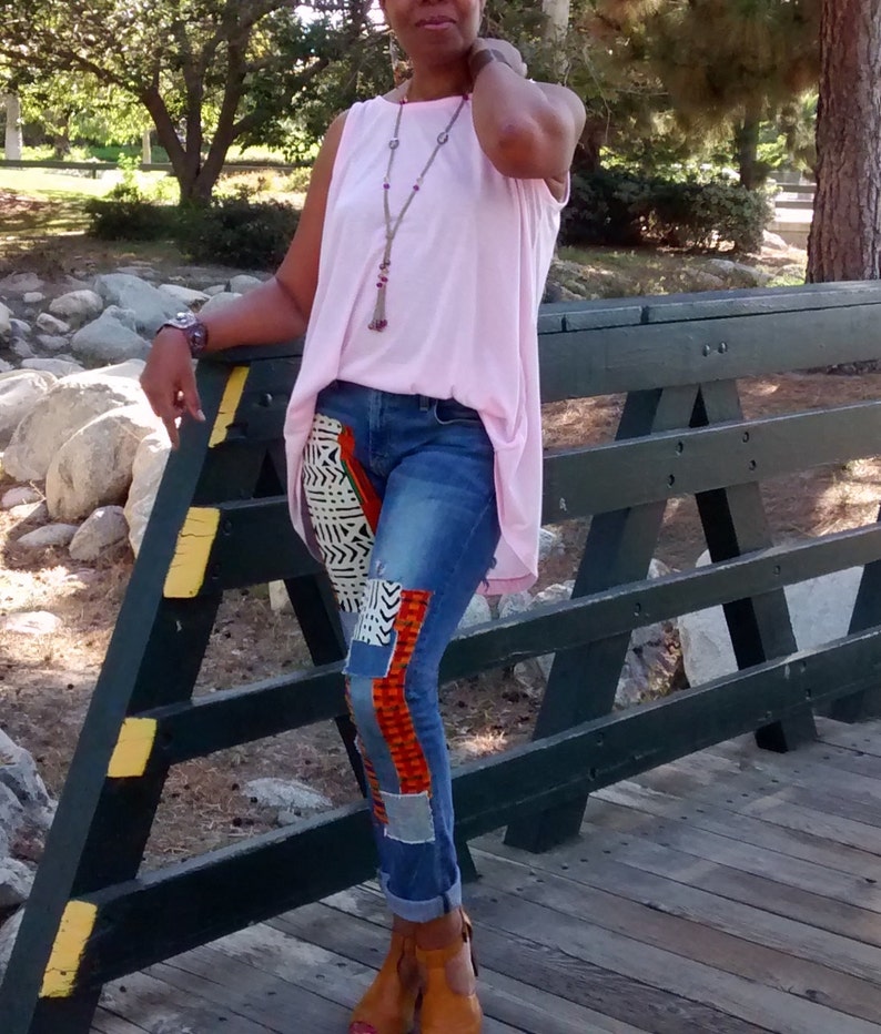 May include: A light pink sleeveless top paired with blue jeans featuring patterned fabric patches on the legs. The outfit is accessorized with a long beaded necklace and brown ankle boots. The person is standing on a wooden bridge.