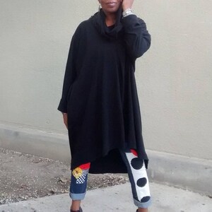 May include: A black, oversized tunic with a cowl neck and long sleeves. The tunic is paired with denim jeans featuring decorative patches and contrasting white and black polka dot accents.