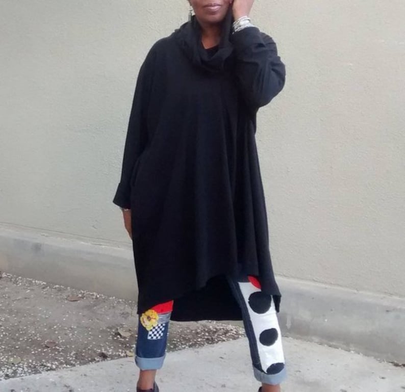 May include: A black, oversized tunic with a cowl neck and long sleeves. The tunic is paired with denim jeans featuring decorative patches and contrasting white and black polka dot accents.