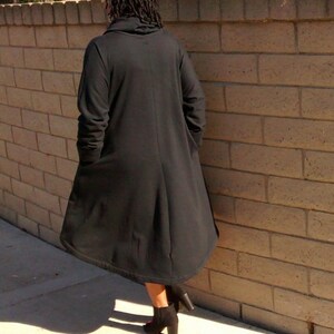 May include: A long, black coat with a high collar and long sleeves. The coat is knee-length and has a flowing, A-line shape. The person is wearing black heeled boots. The background is a brick wall.