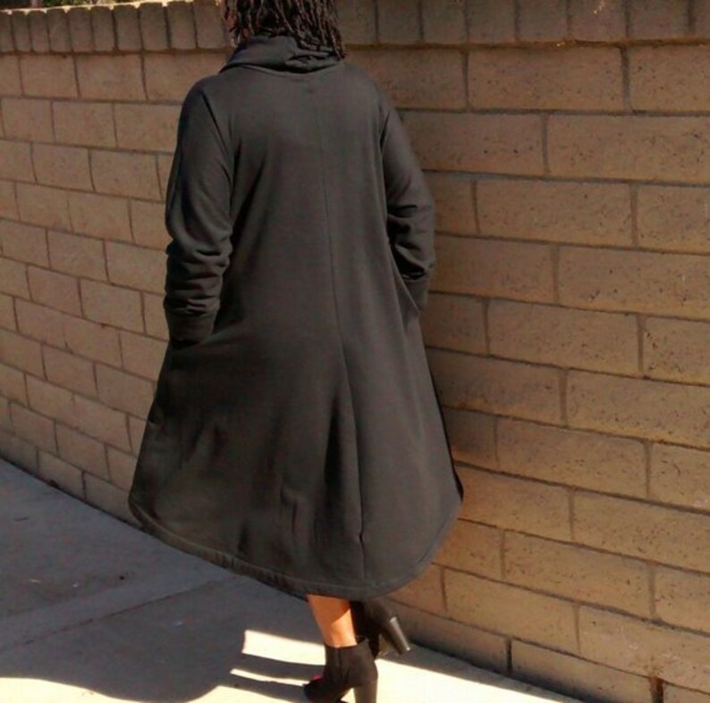 May include: A long, black coat with a high collar and long sleeves. The coat is knee-length and has a flowing, A-line shape. The person is wearing black heeled boots. The background is a brick wall.