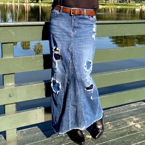 May include: A long denim skirt with distressed detailing. The skirt features ripped sections with visible blue patches, a high waist, and a flared, fishtail-like hem. A brown belt is worn at the waist. The person is wearing black pointed-toe shoes.