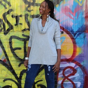 May include: A light gray cowl neck long-sleeve top paired with dark blue distressed jeans. The top has a relaxed fit and a draped neckline. The background features colorful graffiti art on a yellow brick wall.