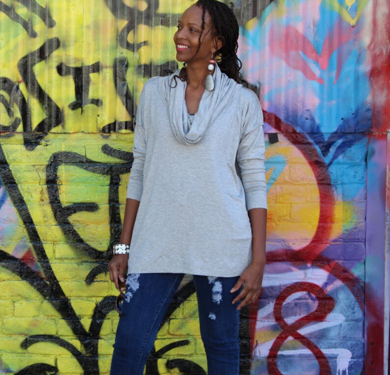 May include: A light gray cowl neck long-sleeve top paired with dark blue distressed jeans. The top has a relaxed fit and a draped neckline. The background features colorful graffiti art on a yellow brick wall.