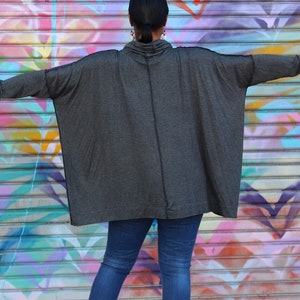 May include: A dark gray, oversized cowl neck top with long sleeves. The top is paired with blue jeans. The background is a colorful graffiti wall. Silver bracelets are visible on the wrists.