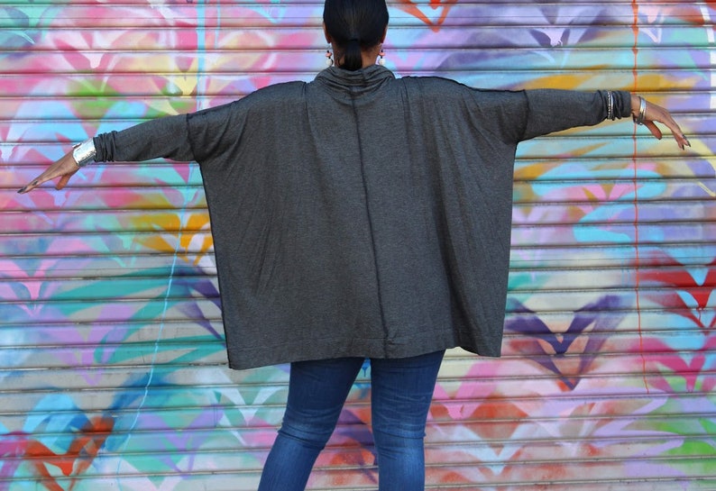 May include: A dark gray, oversized cowl neck top with long sleeves. The top is paired with blue jeans. The background is a colorful graffiti wall. Silver bracelets are visible on the wrists.
