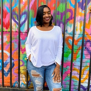 May include: A white long-sleeve shirt with a scoop neck and light blue ripped jeans. The background is a colorful graffiti wall with a black metal fence.