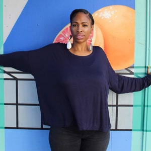 May include: A woman wearing a navy blue long-sleeved top and black pants stands in front of a colorful mural. The mural features geometric shapes, including a large orange fruit. The woman is wearing large, dangling earrings and has yellow nail polish.