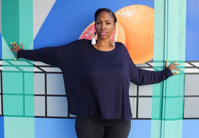May include: A woman wearing a navy blue long-sleeved top and black pants stands in front of a colorful mural. The mural features geometric shapes, including a large orange fruit. The woman is wearing large, dangling earrings and has yellow nail polish.