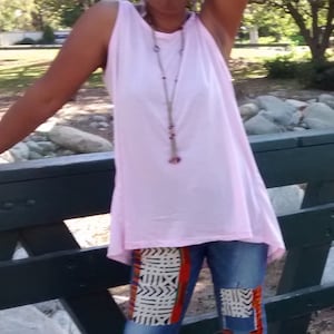 May include: A sleeveless, light pink top paired with blue jeans featuring patterned patches in orange, black, and white. A long necklace with purple accents is visible. The person is standing near a dark green wooden structure.