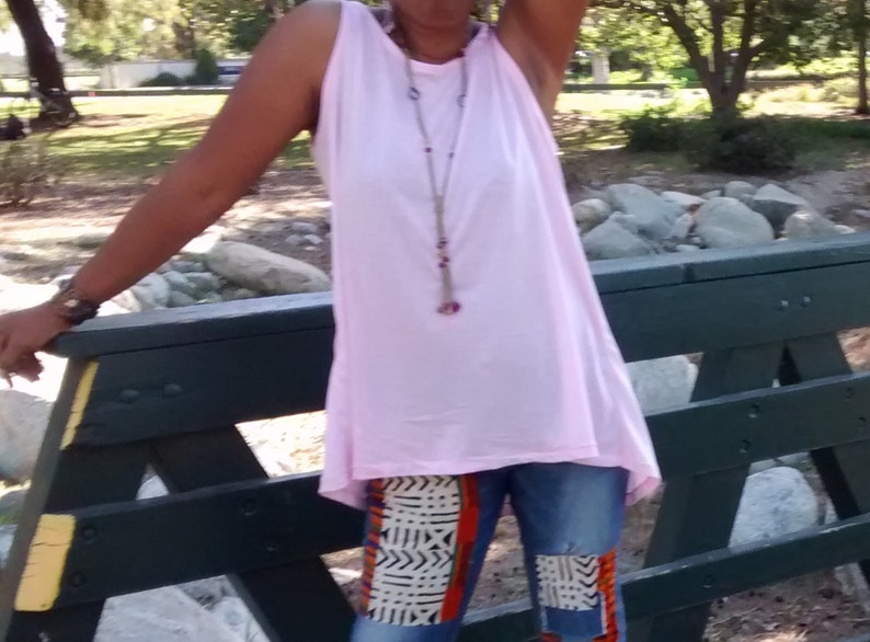 May include: A sleeveless, light pink top paired with blue jeans featuring patterned patches in orange, black, and white. A long necklace with purple accents is visible. The person is standing near a dark green wooden structure.