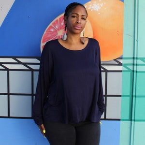 May include: A woman wearing a navy blue long-sleeve top and black pants. The top has a scoop neck and pockets. The background features a colorful mural with a citrus fruit design and geometric shapes.