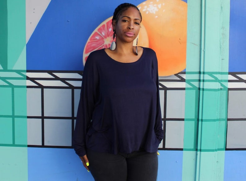 May include: A woman wearing a navy blue long-sleeve top and black pants. The top has a scoop neck and pockets. The background features a colorful mural with a citrus fruit design and geometric shapes.