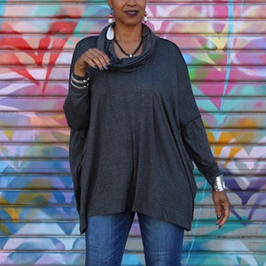 May include: A dark gray cowl neck top with long sleeves. The top has a loose, flowing design. The model is wearing blue jeans and silver bracelets. The background is a colorful graffiti wall.