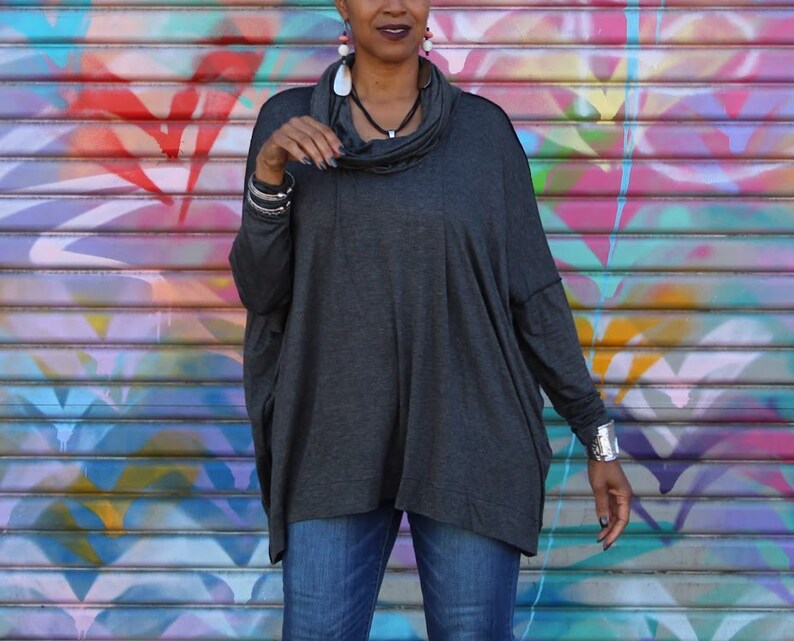 May include: A dark gray cowl neck top with long sleeves. The top has a loose, flowing design. The model is wearing blue jeans and silver bracelets. The background is a colorful graffiti wall.